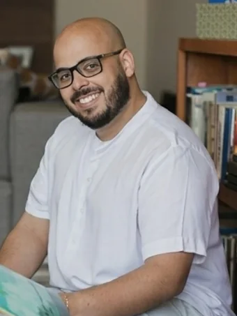 Murilo Vieira, psicólogo e psicanalista, sorrindo, sentado no chão da sala com um livro infantil